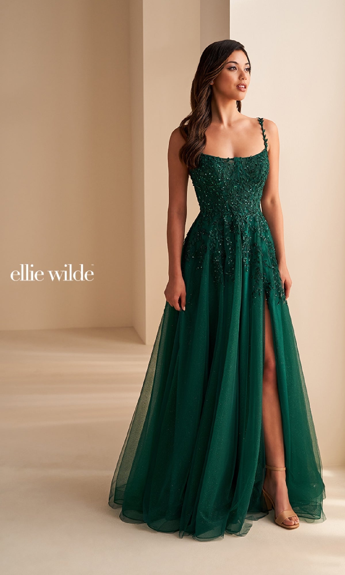 A woman stands against a neutral background in the Ellie Wilde EW36228, a dark green, floor-length prom dress featuring a corset bodice, thin straps, floral embellishments, and a thigh-high slit.